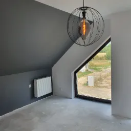 Pokój na poddaszu po malowaniu ścian na szaro i biało, z widocznym grzejnikiem, oknem dachowym z widokiem na zewnątrz i designerską lampą wiszącą z żarówką Edison.