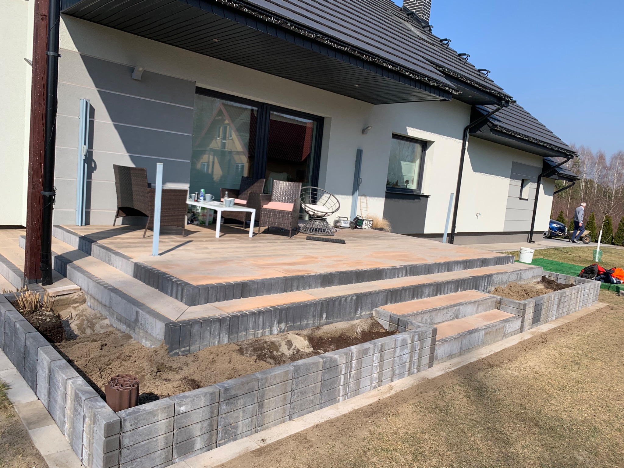 Tarasy kamienne z kostki brukowej i schodami przed domem w trakcie budowy, częściowo obsadzone ziemią, widoczne meble ogrodowe na górnym tarasie.