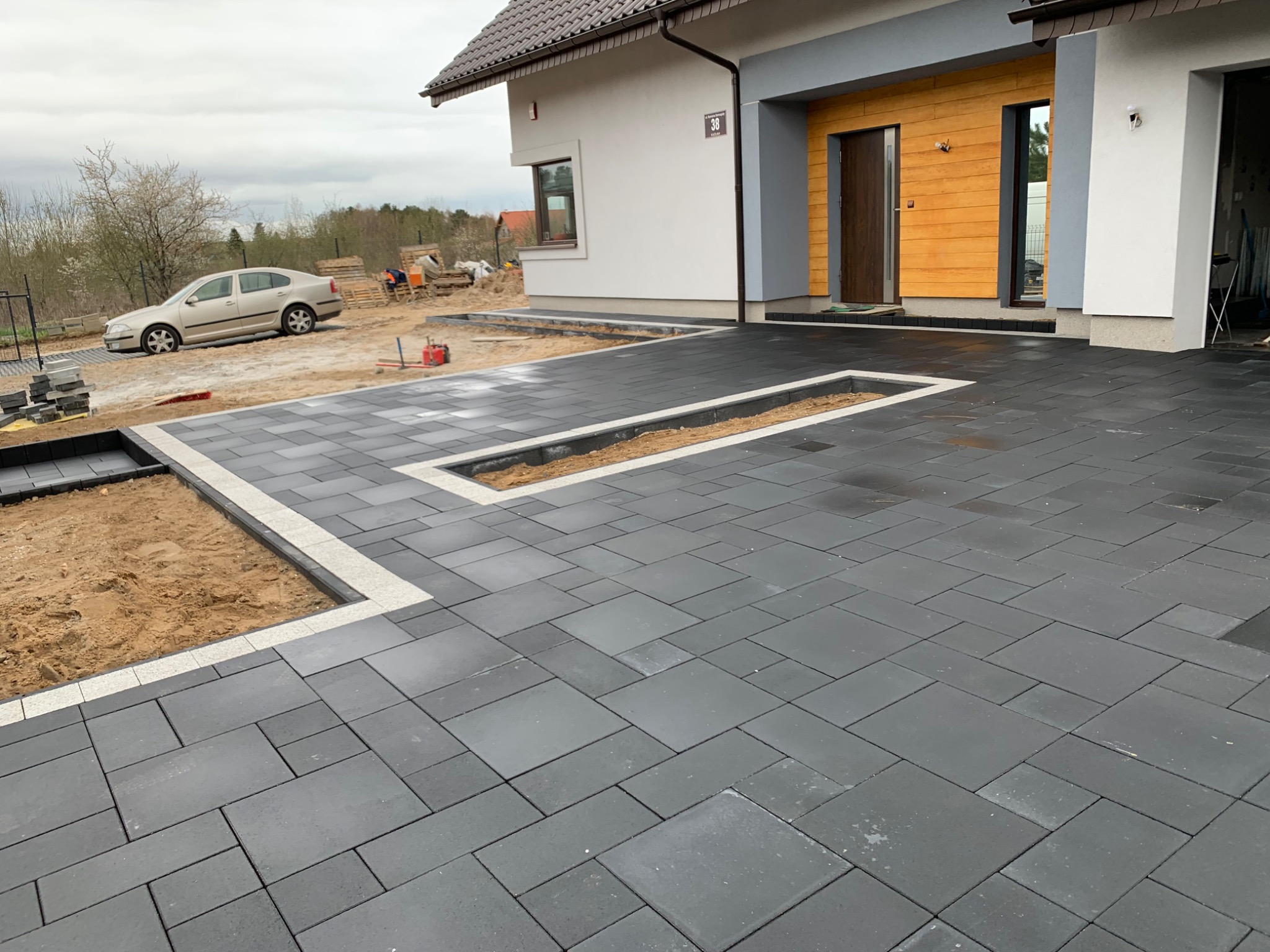 Nowo ułożona, ciemnoszara kostka brukowa na podjeździe przed domem z jasnymi obrzeżami i miejscami na rośliny, widoczny samochód i materiały budowlane w tle.