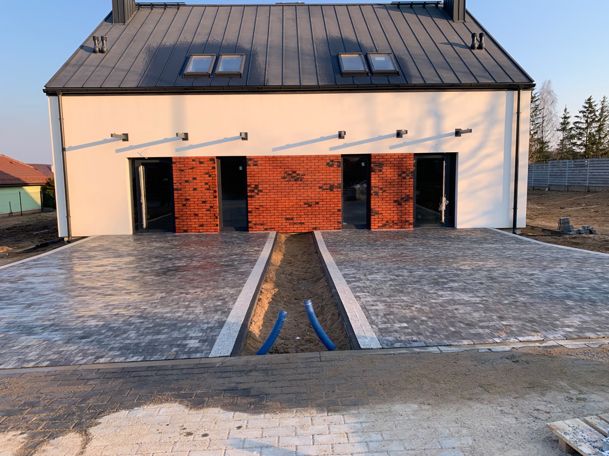 Nowoczesny dom z szarej kostki brukowej ułożonej na podjeździe, z widocznym wykopem na instalacje i fragmentem niezakończonej nawierzchni w tle.
