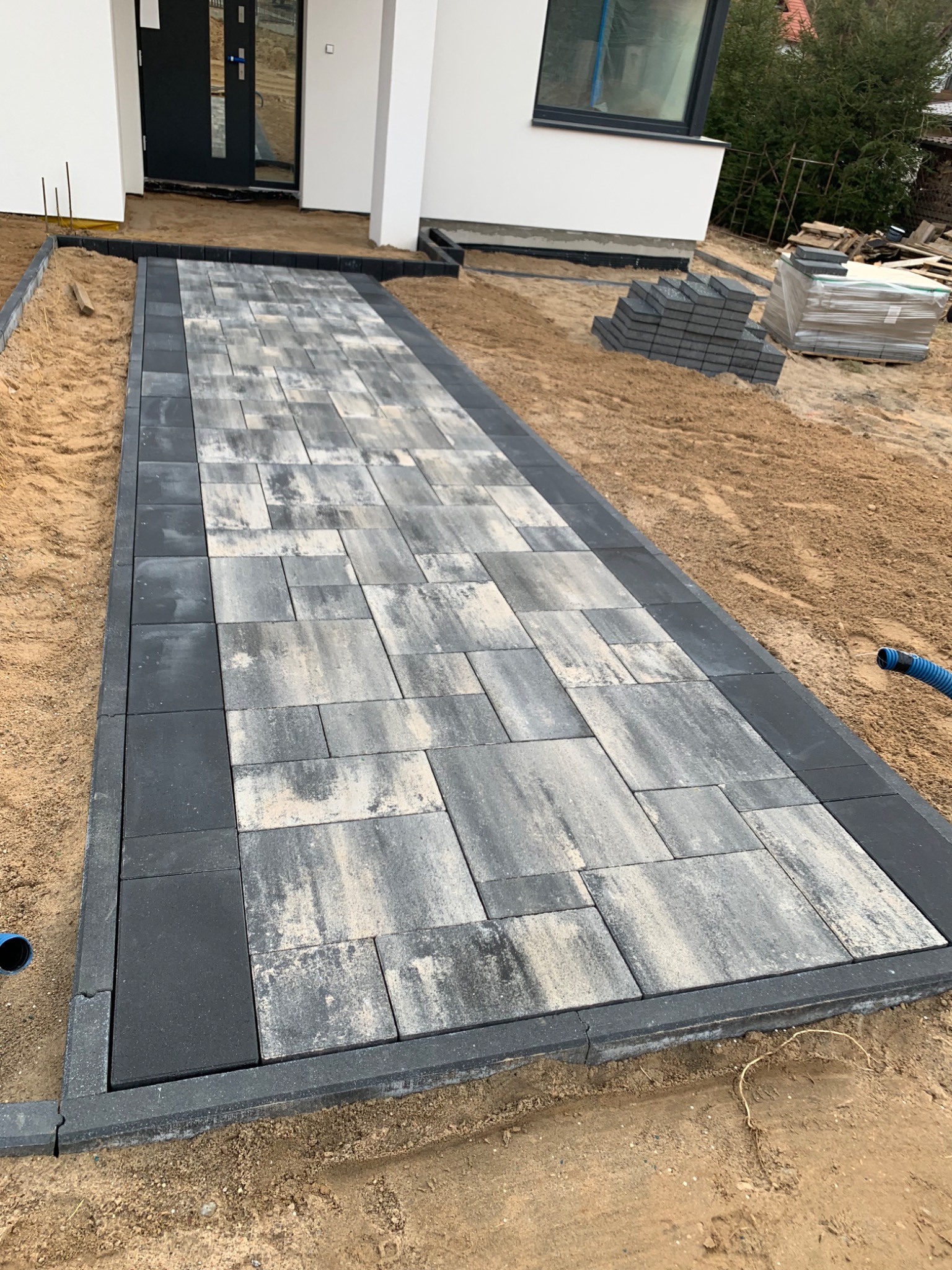 Ścieżka z szarej kostki brukowej prowadząca do nowoczesnego domu, obrzeża z ciemnej kostki, widoczne zapasy kostki na piasku.