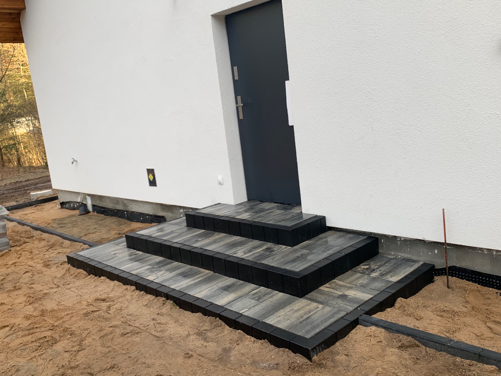 Trzy stopnie schodów wejściowych wyłożone kostką brukową w odcieniach szarości z czarnym obramowaniem, prowadzące do ciemnoszarych drzwi w białym budynku. Widoczny piasek i elementy izolacji przy...