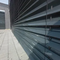 Zewnętrzne żaluzje w kolorze grafitowym, zamontowane na budynku z szarej kostki brukowej, widok z dołu ukazujący strukturę lamel i mechanizm sterowania.