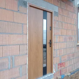 Brązowe drzwi zewnętrzne z czarnym panelem i przeszkleniem, zamontowane w surowej ścianie z cegły ceramicznej, uszczelnione pianką montażową.