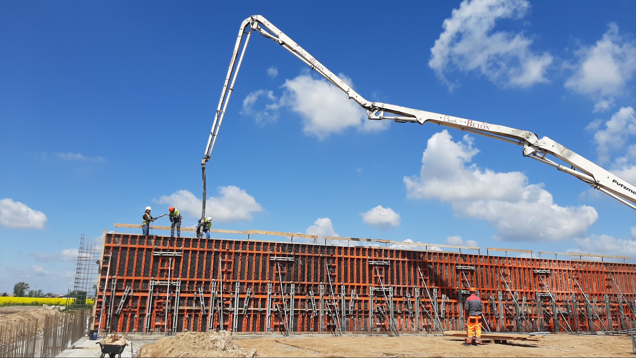Betonowanie ściany szalunkowej przy użyciu pompy do betonu, pracownicy w kaskach nadzorują proces, w tle błękitne niebo z chmurami.