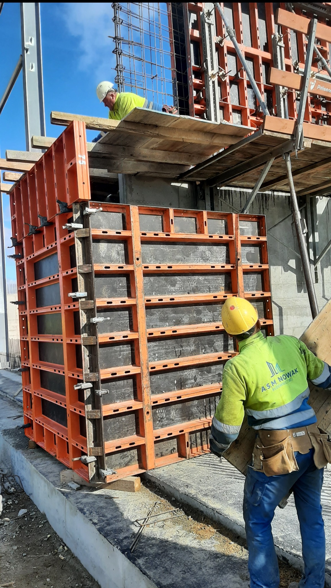 Montaż szalunków systemowych z pomarańczowymi ramami i czarnymi panelami przez dwóch pracowników w kaskach i roboczych ubraniach, z widocznym zbrojeniem w tle, na placu budowy.