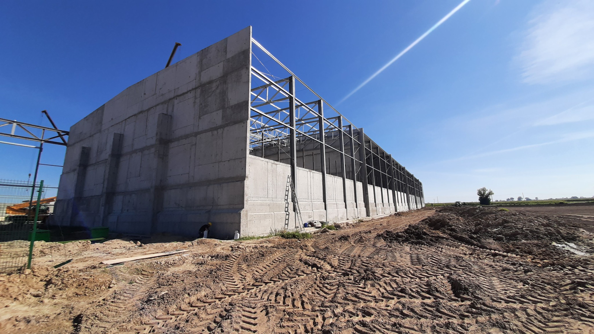 Surowy budynek żelbetowy w trakcie budowy, widoczne szalunki i konstrukcja stalowa dachu na tle błękitnego nieba, ziemia wokół budynku z wyraźnymi śladami opon.