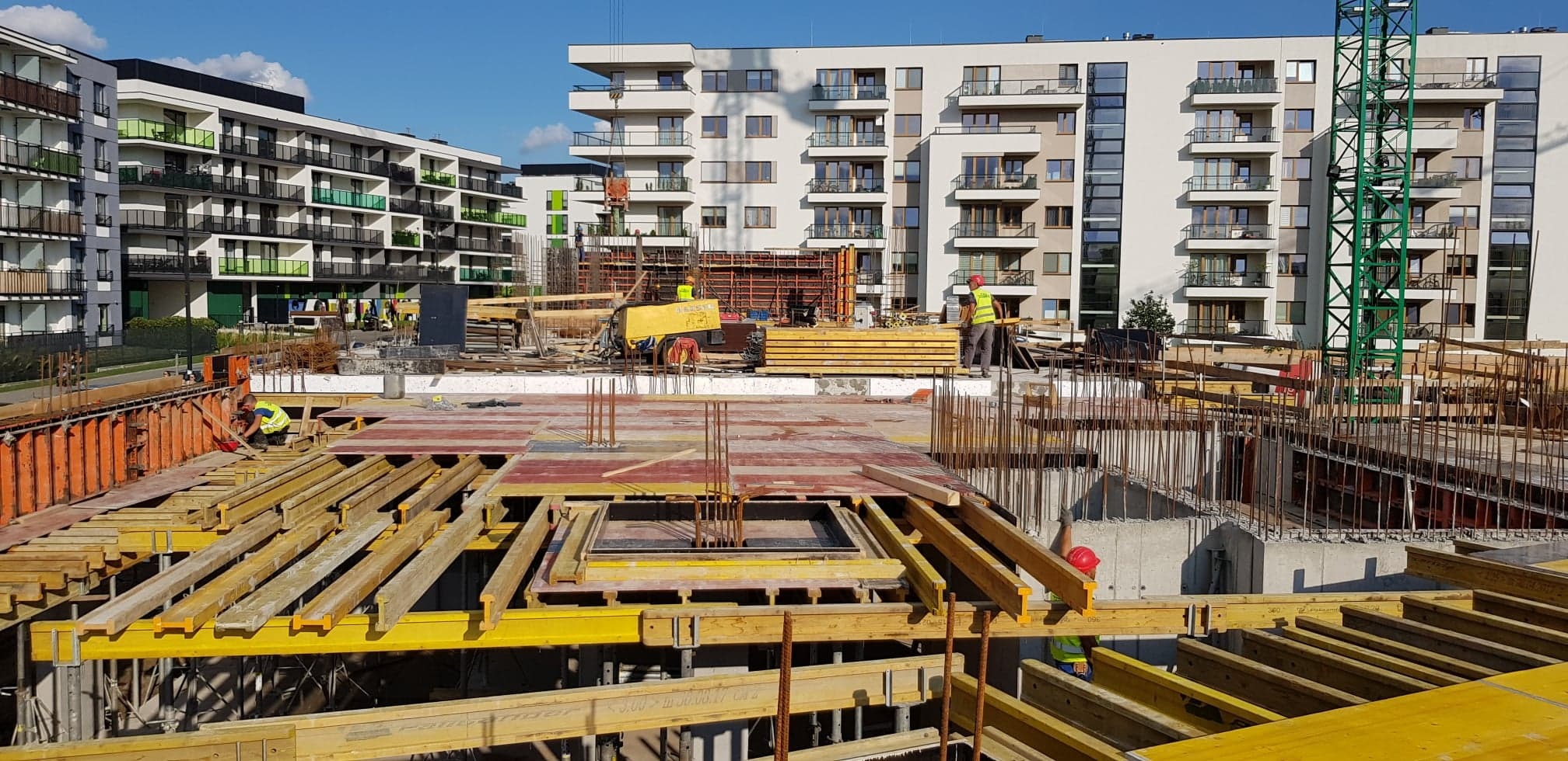 Budowa budynku wielorodzinnego w trakcie realizacji: widoczne szalunki, zbrojenia, pracownicy w kamizelkach odblaskowych i kaskach, dźwig budowlany w tle.