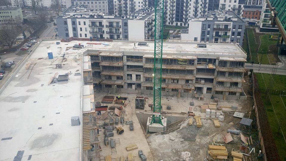 Wysoki kąt widokowy na plac budowy wielopiętrowego budynku z żelbetową konstrukcją, z widocznym żurawiem wieżowym w centrum kadru oraz elementami szalunków i zbrojeń.