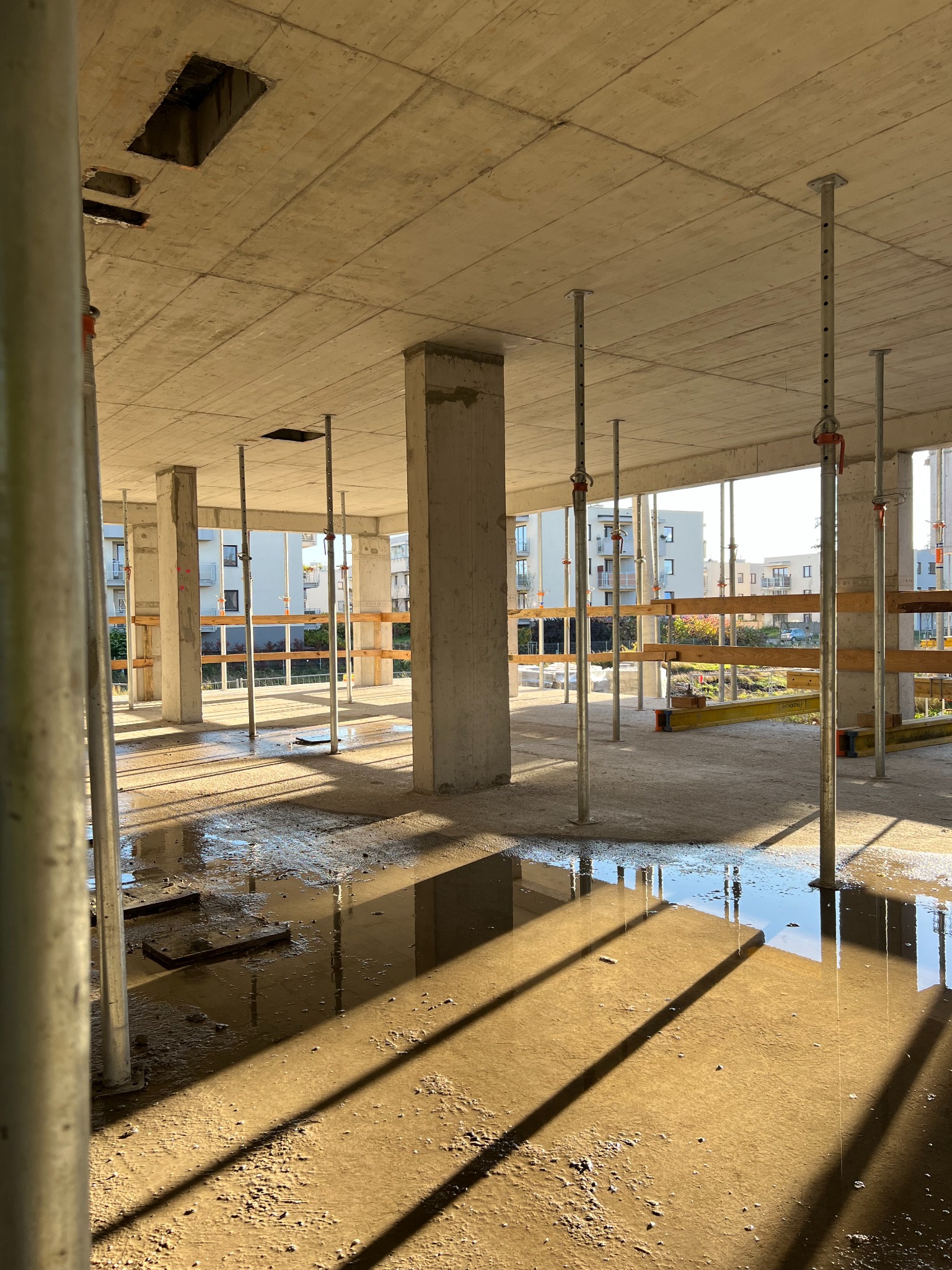 Surowy stan budynku żelbetowego w trakcie budowy, widoczne mokre podłoże, betonowe słupy, strop z otworami, podpory stalowe i drewniane szalunki.