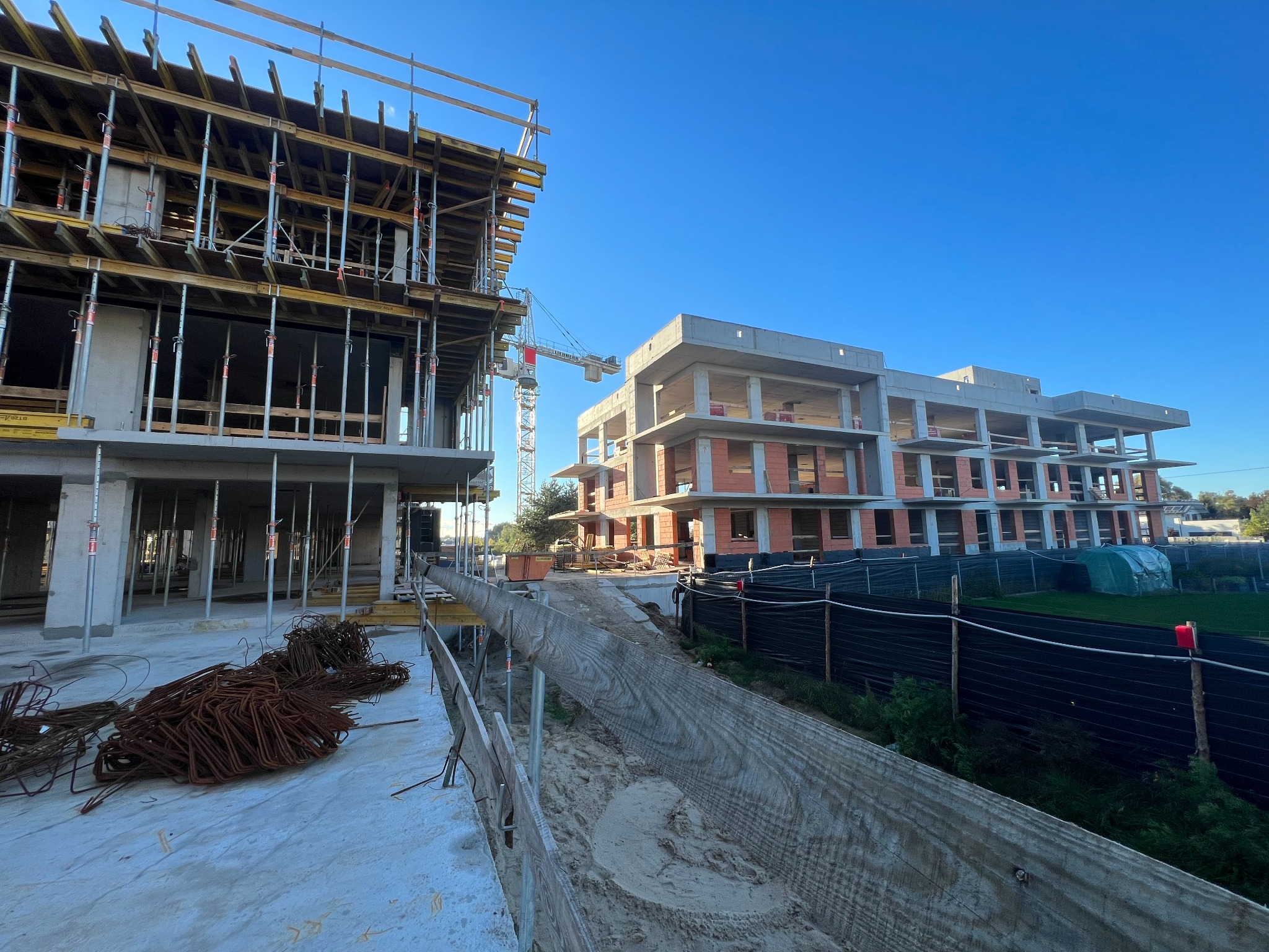 Budowa wielorodzinnego budynku żelbetowego w trakcie realizacji, widoczne szalunki, rusztowania i składowane pręty zbrojeniowe, w tle żuraw budowlany i częściowo wykończony budynek z czerwonej cegły.