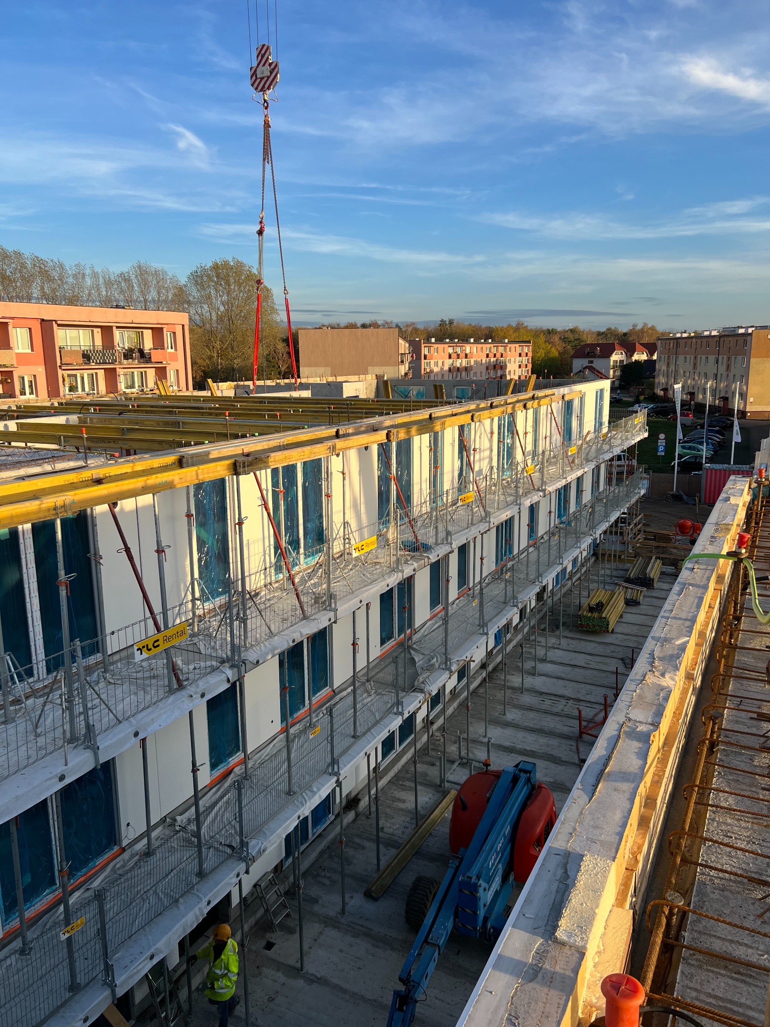 Wielopiętrowy budynek modułowy w trakcie montażu, widoczny dźwig z hakiem, robotnik w żółtym kasku i podnośnik koszowy na placu budowy, w tle zabudowa mieszkalna.