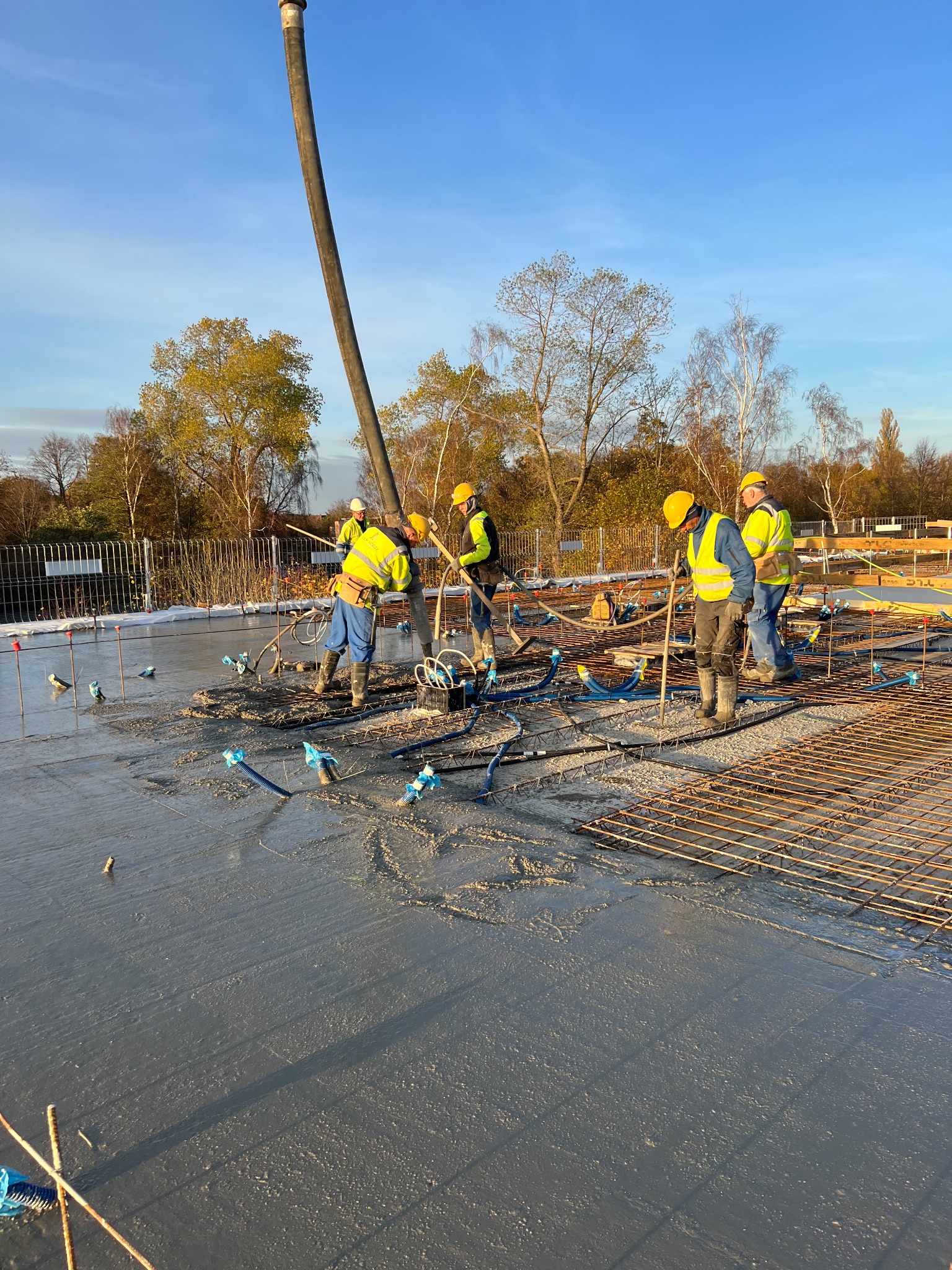 Wylewanie betonu na strop zbrojony, widoczni robotnicy w żółtych kaskach i kamizelkach odblaskowych, niebieskie rurki instalacyjne wystające z betonu, w tle drzewa i błękitne niebo.