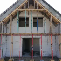 Wznoszenie ścian domu z betonu komórkowego, widoczne otwory okienne, drewniana konstrukcja dachu i podpory szalunkowe w kolorach czerwonym, żółtym i niebieskim.