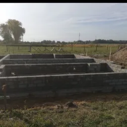 Wznoszenie fundamentów z bloczków betonowych na terenie budowy, widoczne elementy szalunku i hałda ziemi w tle.