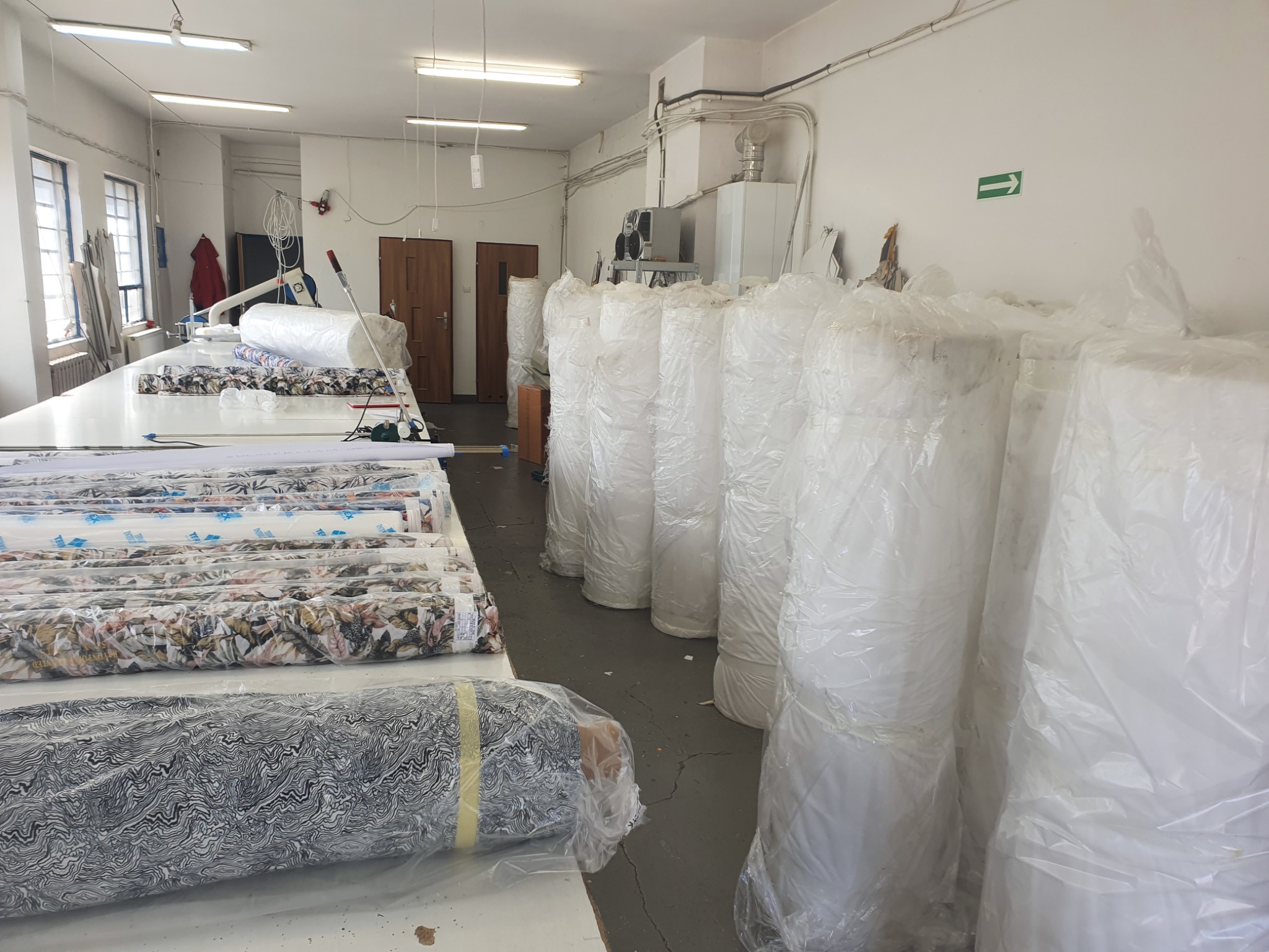 Wnętrze hali z długim stołem roboczym wypełnionym rolkami materiałów w różnych wzorach, obok stos rolek materiałów zapakowanych w przezroczystą folię.