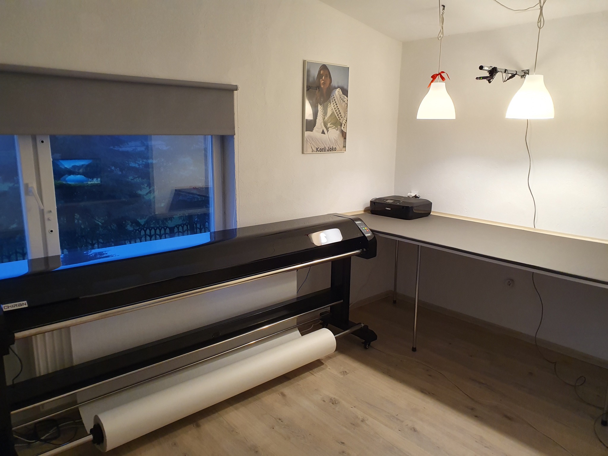 Szerokoformatowa drukarka z rolką papieru w pracowni, widok na okno z roletą i plakat na ścianie.