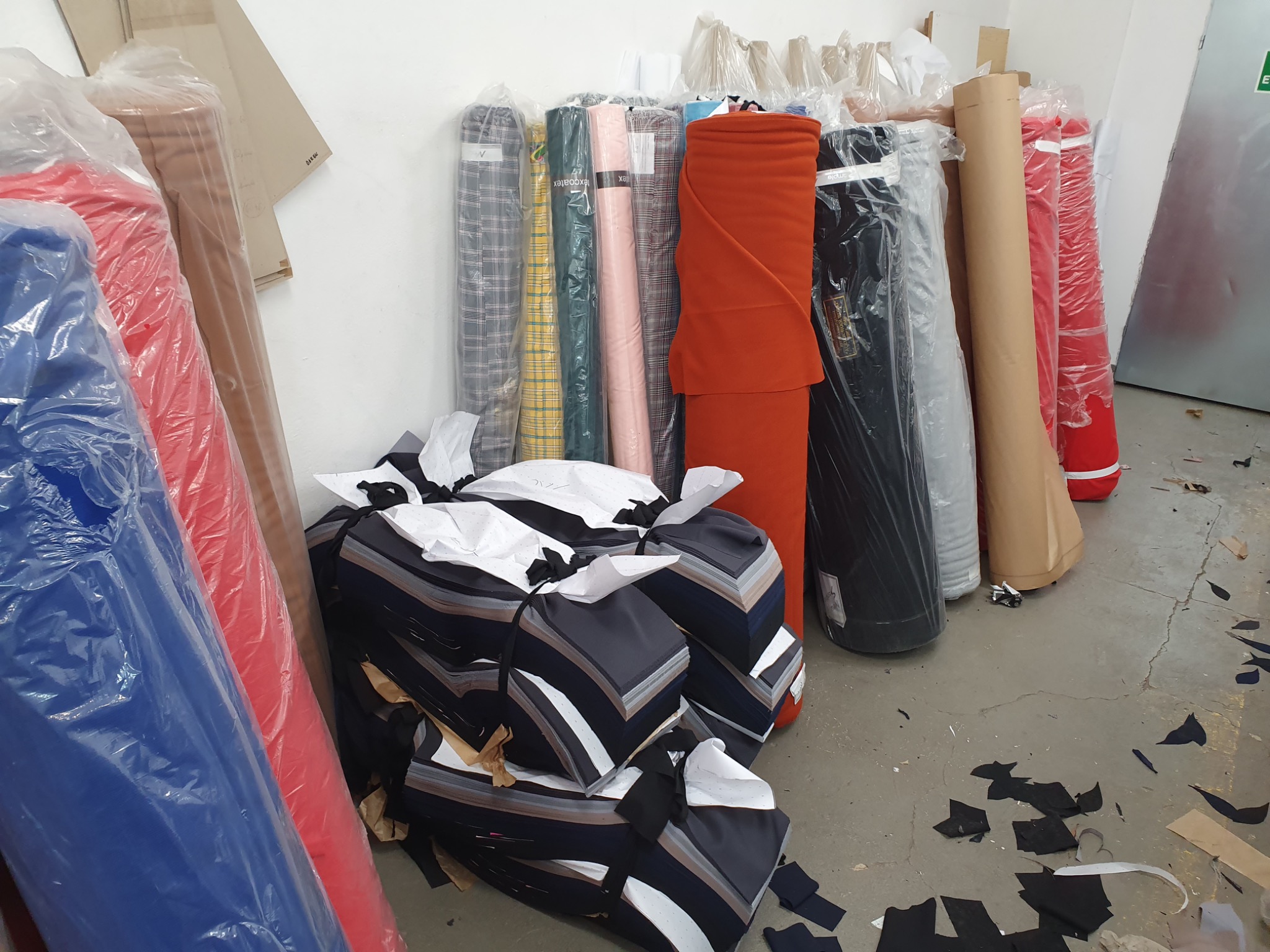 Skladowane rolki różnokolorowych materiałów tekstylnych, niektóre zapakowane w folię, oraz stosy wykrojonych elementów odzieży z ciemnego materiału w pomieszczeniu produkcyjnym.