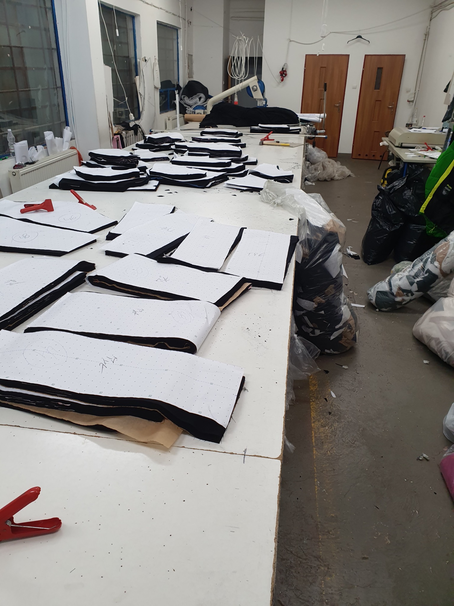 Stół w pracowni z rozłożonymi warstwami tkaniny, na których leżą papierowe wykroje z naniesionymi numerami, czerwone klamry i nożyczki krawieckie w tle, worki z resztkami materiałów obok.