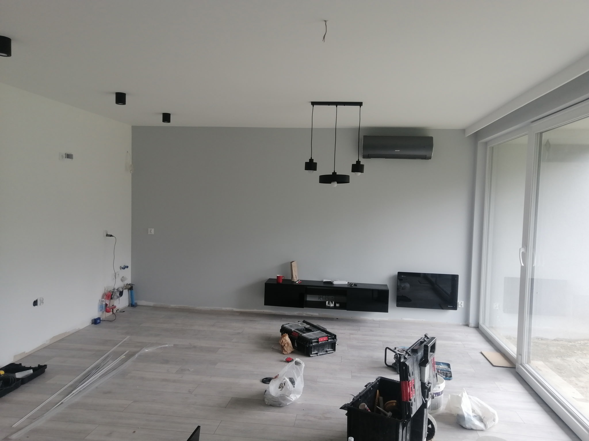 Jasne wnętrze w trakcie wykańczania: szare ściany, panele podłogowe, wisząca szafka RTV, klimatyzator i narzędzia glazurnicze. Widoczne przygotowania do dalszych prac.
