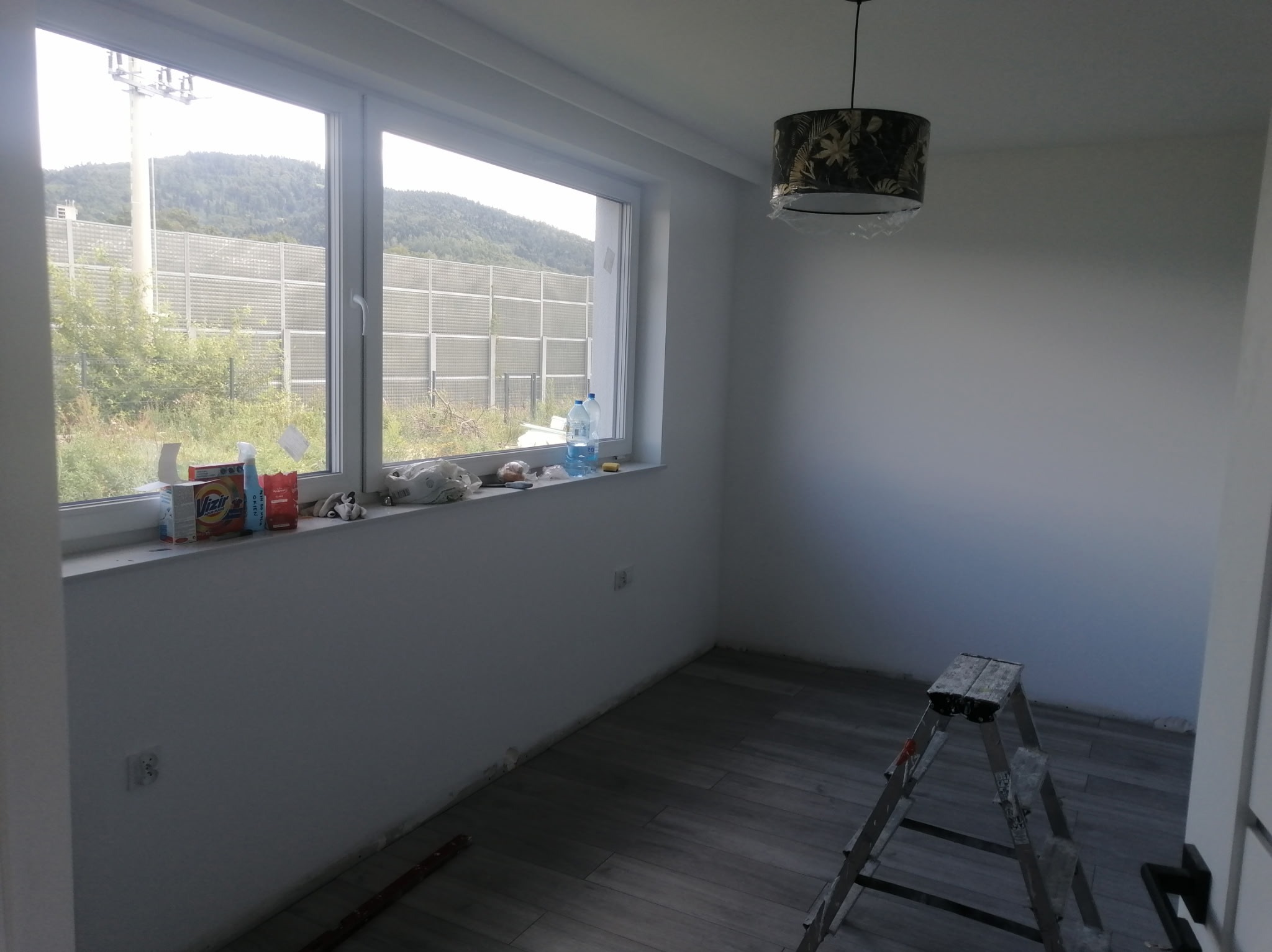 Wnętrze pokoju po malowaniu: białe ściany, szare panele podłogowe, drabina malarska. Na parapecie okna widoczne akcesoria malarskie i butelki z wodą. Widok na zewnątrz.