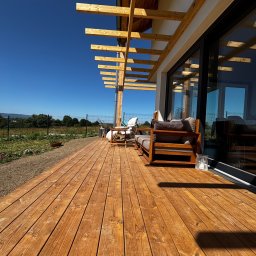 Spod Hebla - Drewniany taras z jasnymi deskami i zadaszeniem z belek. Widoczne meble ogrodowe i przesuwne okno. W tle zieleń i błękitne niebo.