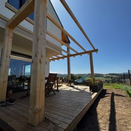 Spod Hebla - Drewniany taras w trakcie budowy, widok na krajobraz. Elementy konstrukcyjne z surowego drewna, deski tarasowe, krzesła i hamak w tle. Słoneczny dzień.