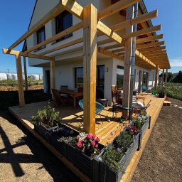 Spod Hebla - Nowoczesny taras z drewna z pergolą i donicami z kwiatami, przylegający do białego domu. Widok na ogród i błękitne niebo w słoneczny dzień.