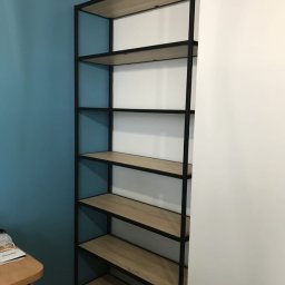 Spod Hebla - Metalowy regał z drewnianymi półkami na tle niebieskiej i białej ściany. Prosta konstrukcja, widoczne połączenia elementów. Minimalistyczny design.