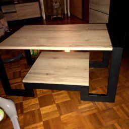 Spod Hebla - Stolik kawowy z blatem drewnopodobnym i czarnymi, metalowymi nogami, na tle parkietu. Widoczna dolna półka i fragmenty otoczenia.