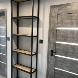 Spod Hebla - Wysoki regał z drewnianymi półkami i czarną, metalową ramą, stojący obok nowoczesnych drzwi z szarego betonu z podłużnymi, białymi paskami.