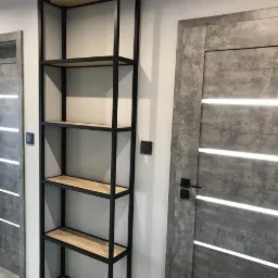 Wysoki regał z drewnianymi półkami i czarną, metalową ramą, stojący obok nowoczesnych drzwi z szarego betonu z podłużnymi, białymi paskami.