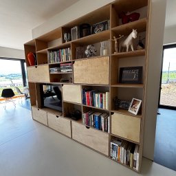 Spod Hebla - Półka z jasnej sklejki, wbudowana w ścianę, z licznymi przegródkami, książkami i dekoracjami. W środkowej części wnęka na telewizor. Nowoczesny minimalizm.