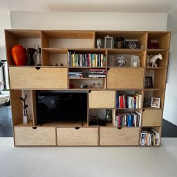 Spod Hebla - Duża, drewniana szafa z półkami i szufladami z jasnej sklejki, w której znajduje się telewizor, książki i dekoracje. Szafa stoi na tle białej ściany.