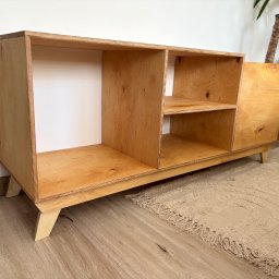 Spod Hebla - Drewniana komoda z półkami z jasnej sklejki, na niskich nóżkach, w minimalistycznym stylu. W tle widoczny dywan i roślina doniczkowa.