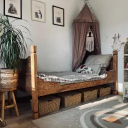 Drewniane łóżko dziecięce z baldachimem w pokoju, z trzema koszami pod spodem. Na ścianach wiszą obrazki i naklejki. Obok stoi regał z książkami.