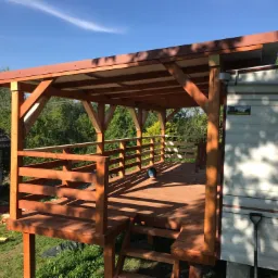 Nowa, drewniana altana z brązowym dachem i balustradą, częściowo ukończona, z widocznymi schodami wejściowymi i otoczeniem zieleni oraz przyczepą kempingową w tle.