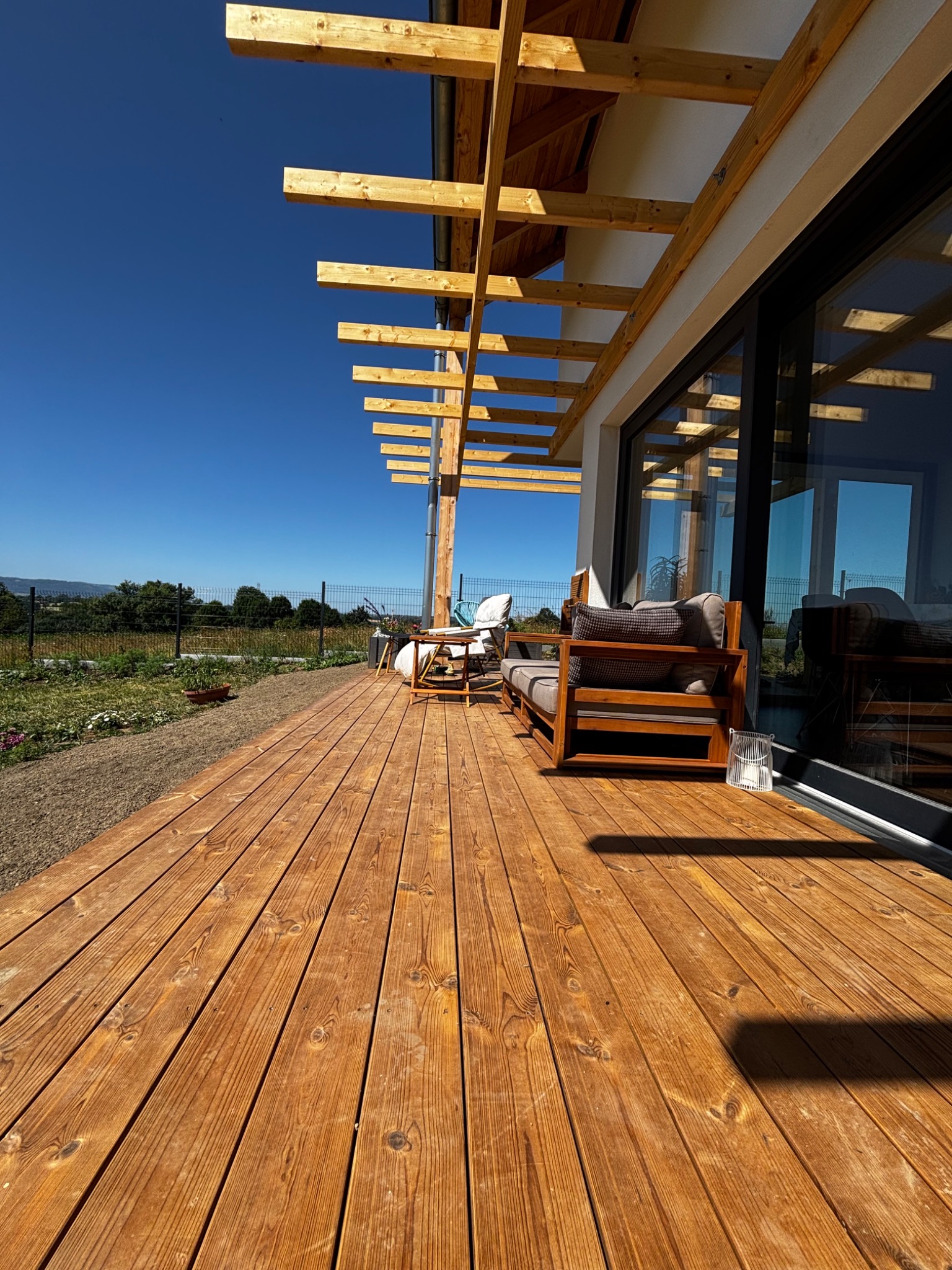 Drewniany taras z jasnymi deskami i zadaszeniem z belek. Widoczne meble ogrodowe i przesuwne okno. W tle zieleń i błękitne niebo.