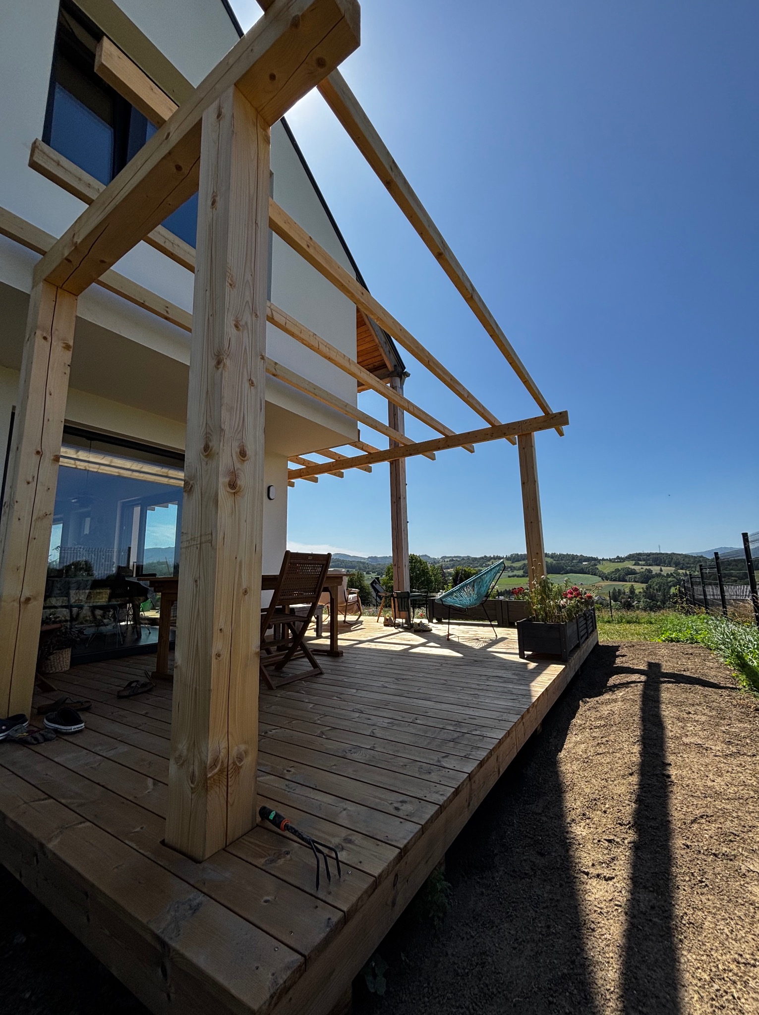 Drewniany taras w trakcie budowy, widok na krajobraz. Elementy konstrukcyjne z surowego drewna, deski tarasowe, krzesła i hamak w tle. Słoneczny dzień.
