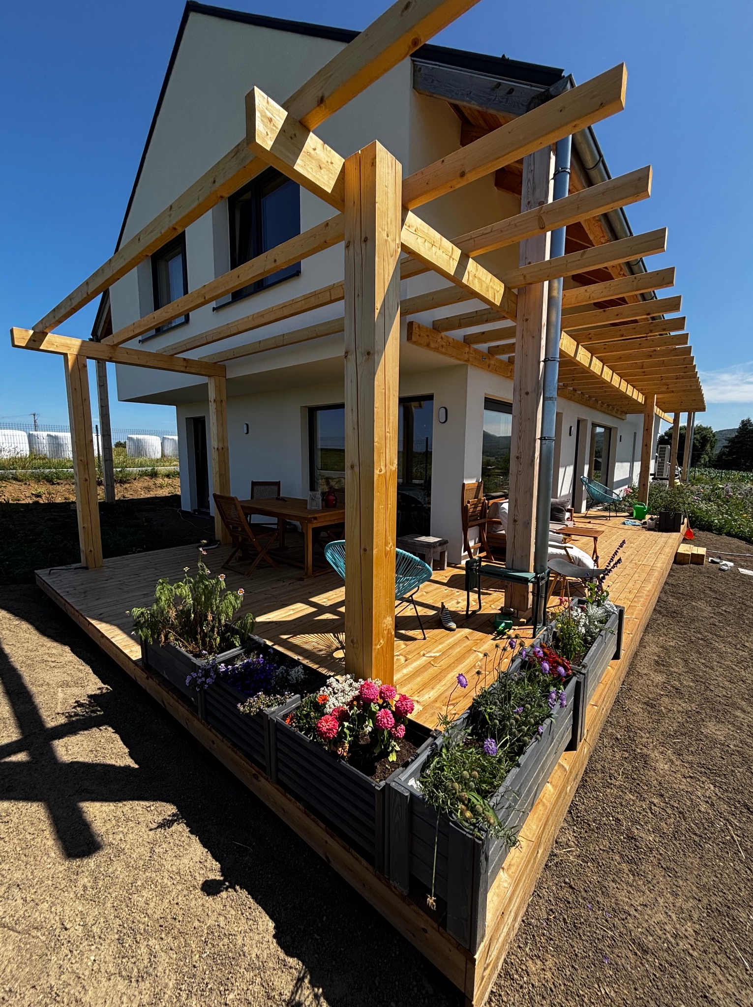 Nowoczesny taras z drewna z pergolą i donicami z kwiatami, przylegający do białego domu. Widok na ogród i błękitne niebo w słoneczny dzień.