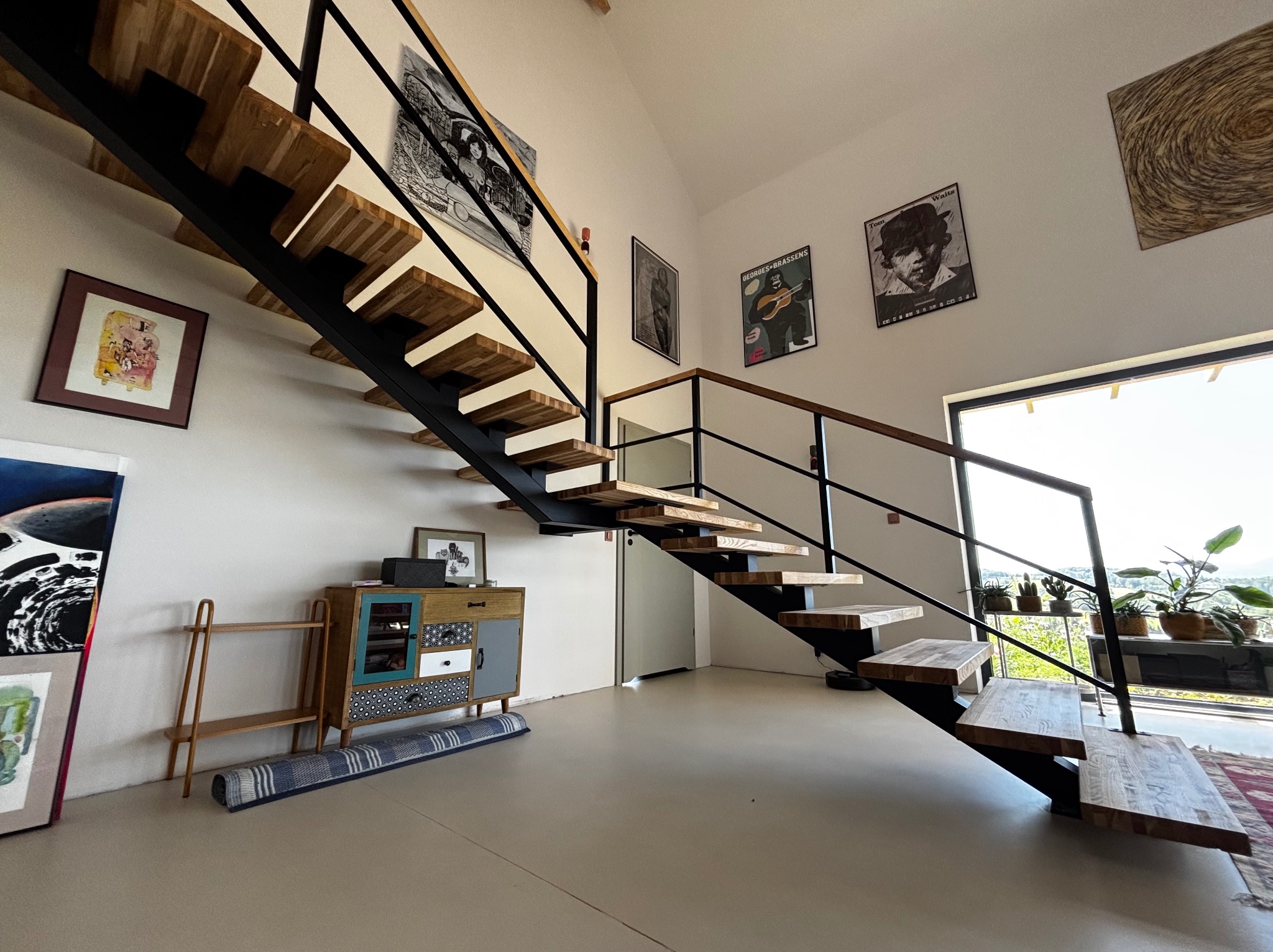 Nowoczesne schody drewniane z czarną metalową konstrukcją w jasnym wnętrzu z dekoracjami ściennymi i roślinami w tle. Minimalistyczny design.