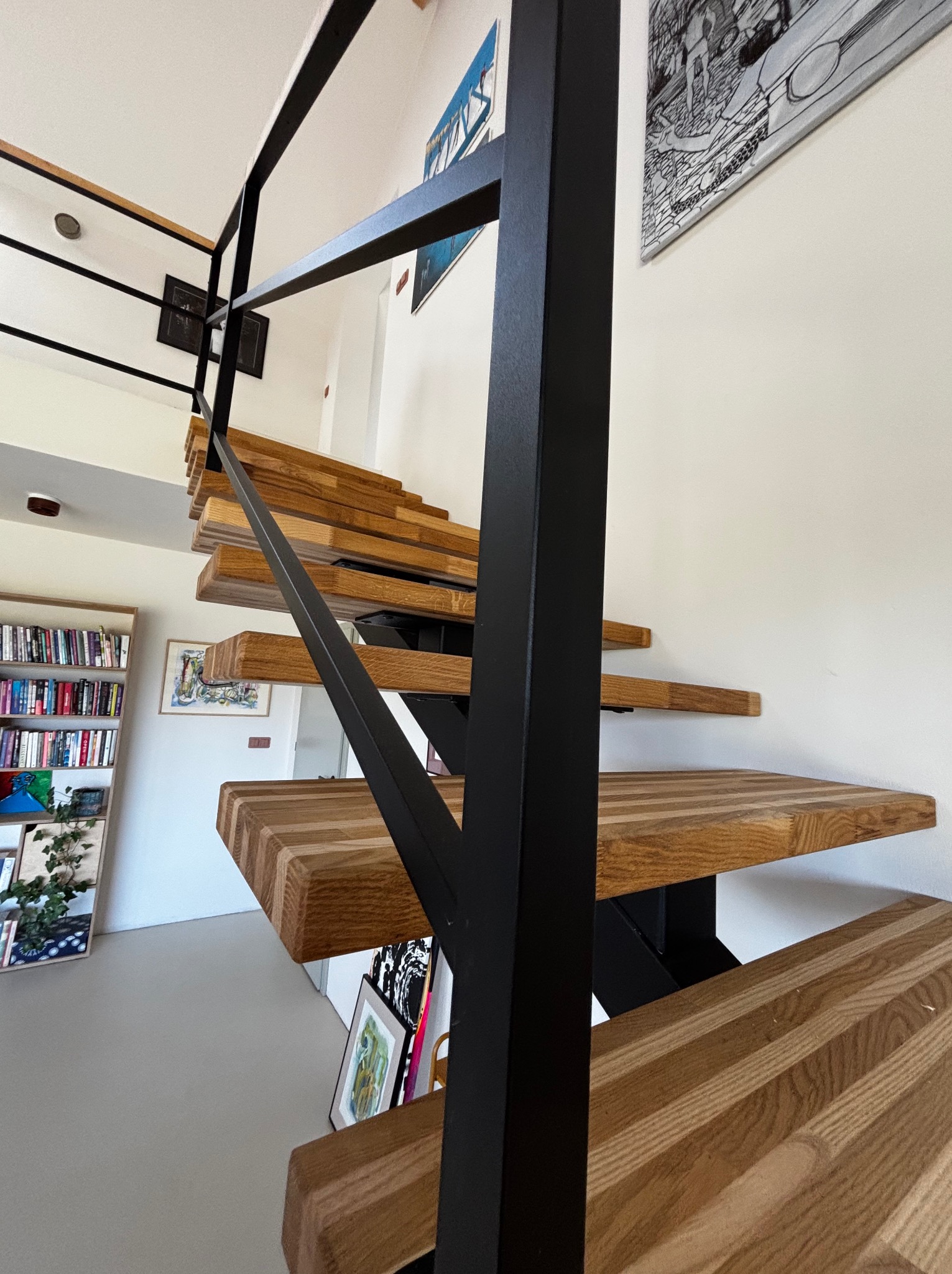 Nowoczesne, drewniane schody z czarną metalową konstrukcją. Stopnie z wyraźnym usłojeniem. Widok z dołu, fragment biblioteczki w tle.