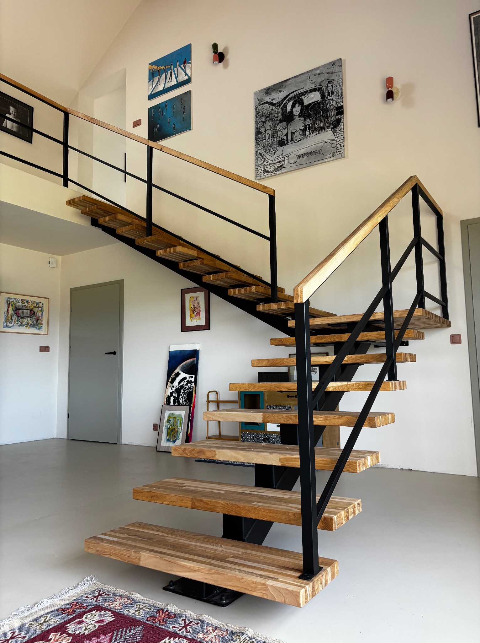 Nowoczesne schody drewniane z czarną balustradą w jasnym wnętrzu. Stopnie z litego drewna, minimalistyczny design, obrazy na ścianie, dywanik na podłodze.