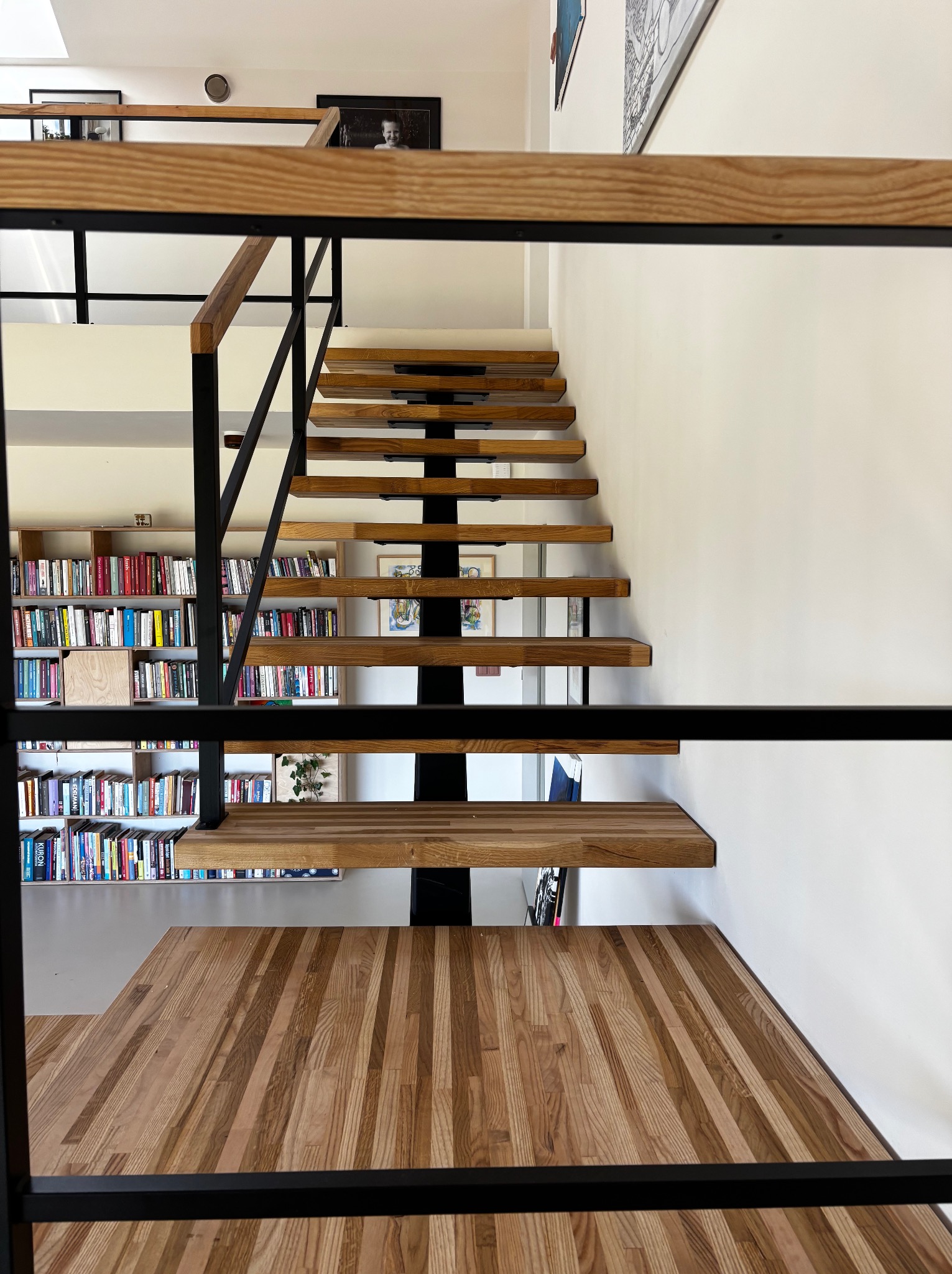 Nowoczesne schody drewniane z czarną metalową konstrukcją, widok z dołu, z biblioteczką w tle, jasne oświetlenie.