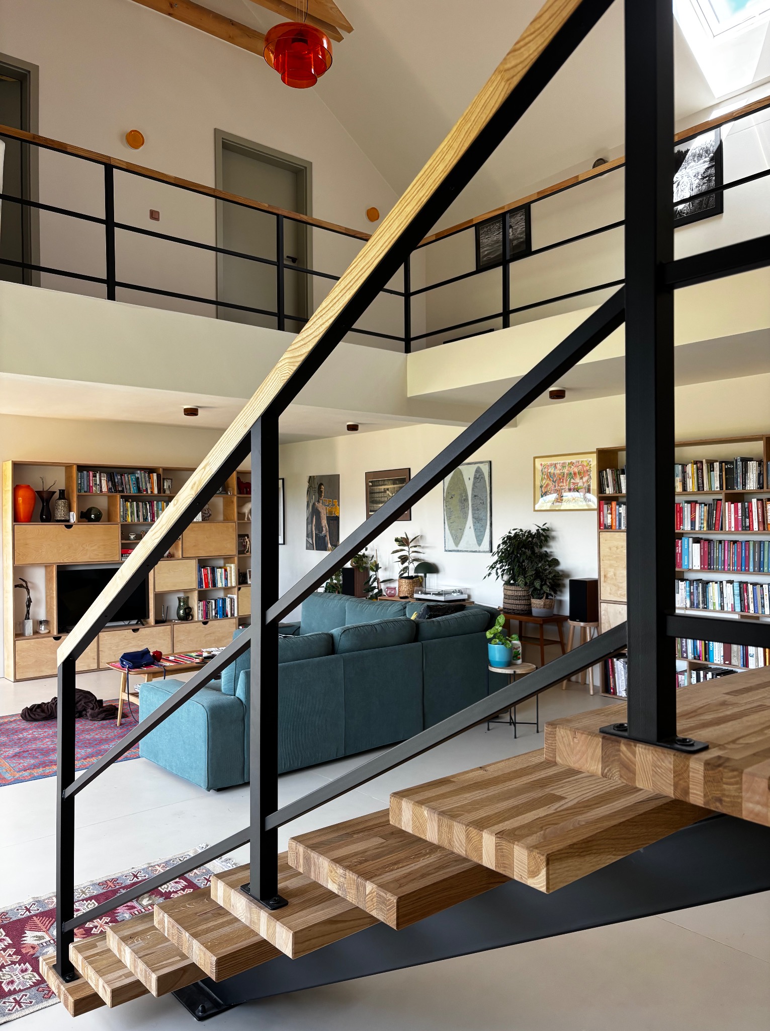 Nowoczesne schody drewniane z czarną balustradą, widoczne z dołu, prowadzące na antresolę w jasnym, przestronnym salonie z biblioteczką i turkusową sofą.