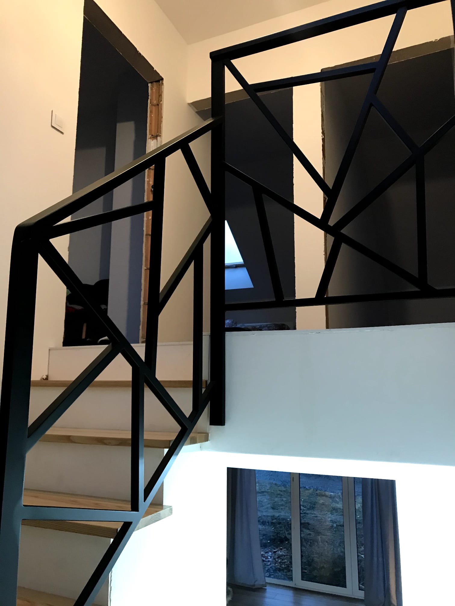 Nowoczesne schody drewniane z czarną, geometryczną balustradą. Widok z dołu, na pierwszym planie stopnie i balustrada, w tle okno i widok na zewnątrz.