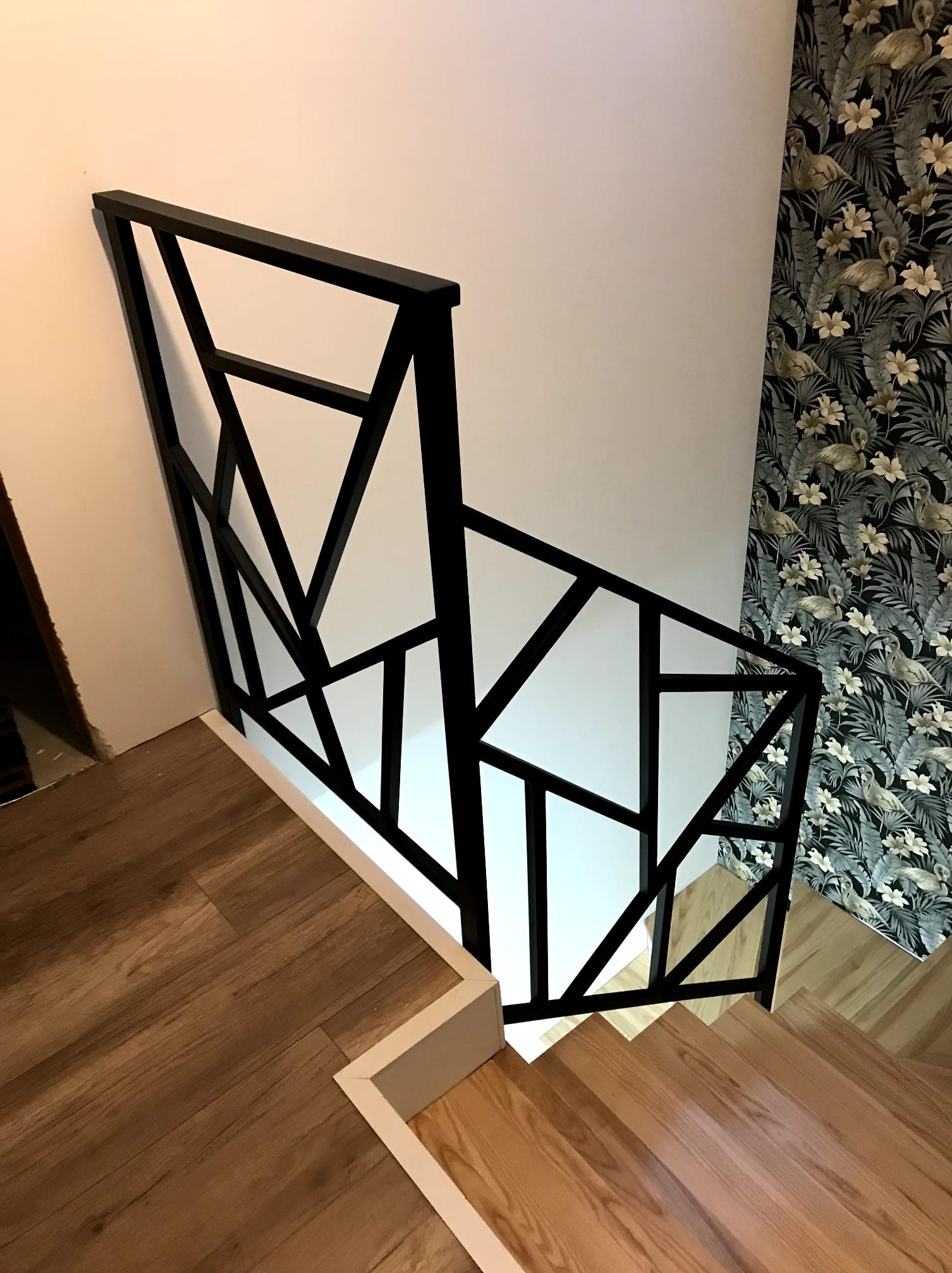 Nowoczesna balustrada schodowa z czarnych, geometrycznych elementów w Bielsko-Białej. Drewniane schody i ściana z wzorzystą tapetą tworzą stylowe wnętrze.