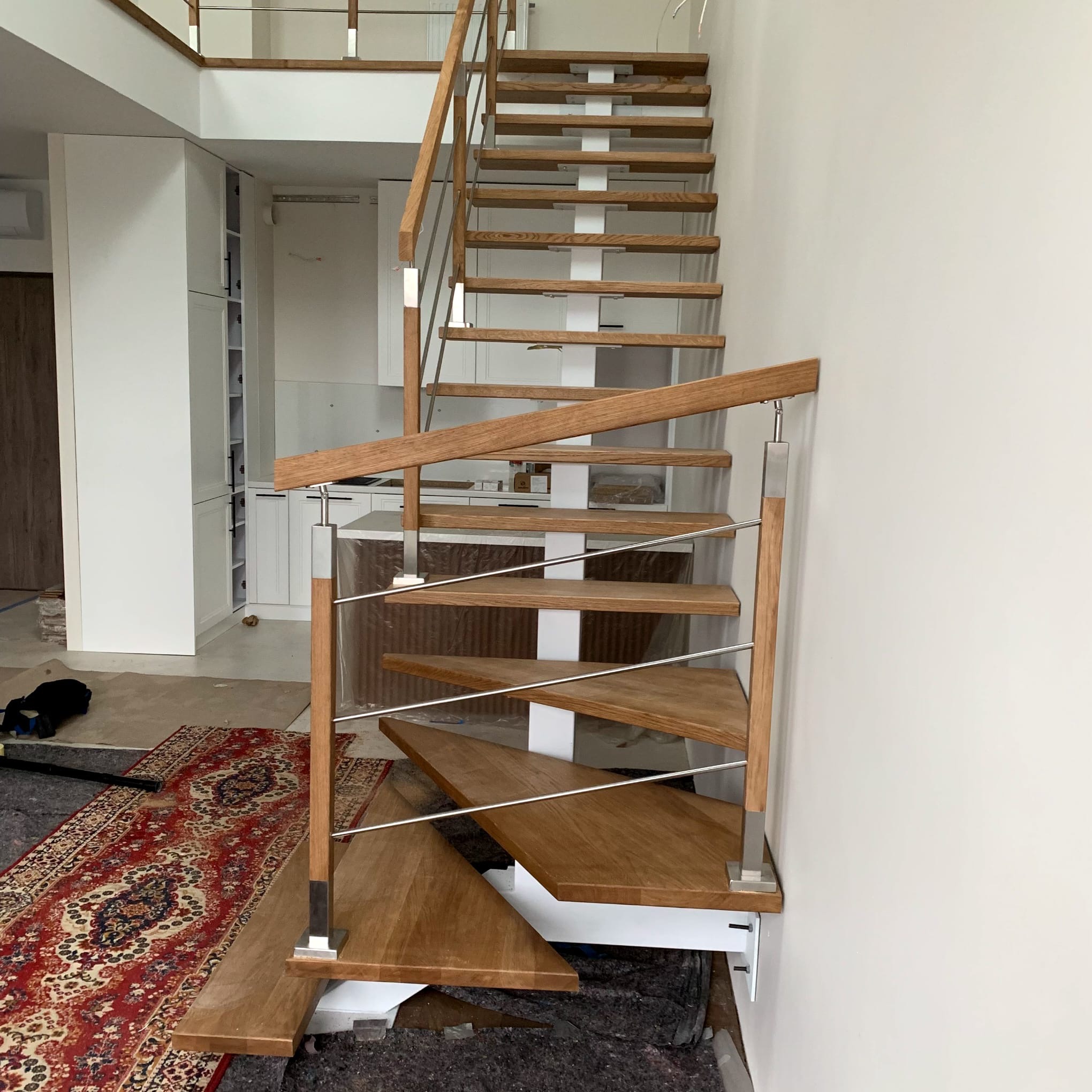 Drewniane schody z metalowymi elementami, widok z dołu, w tle biała zabudowa kuchenna. Wykończenie schodów w jasnym odcieniu drewna.