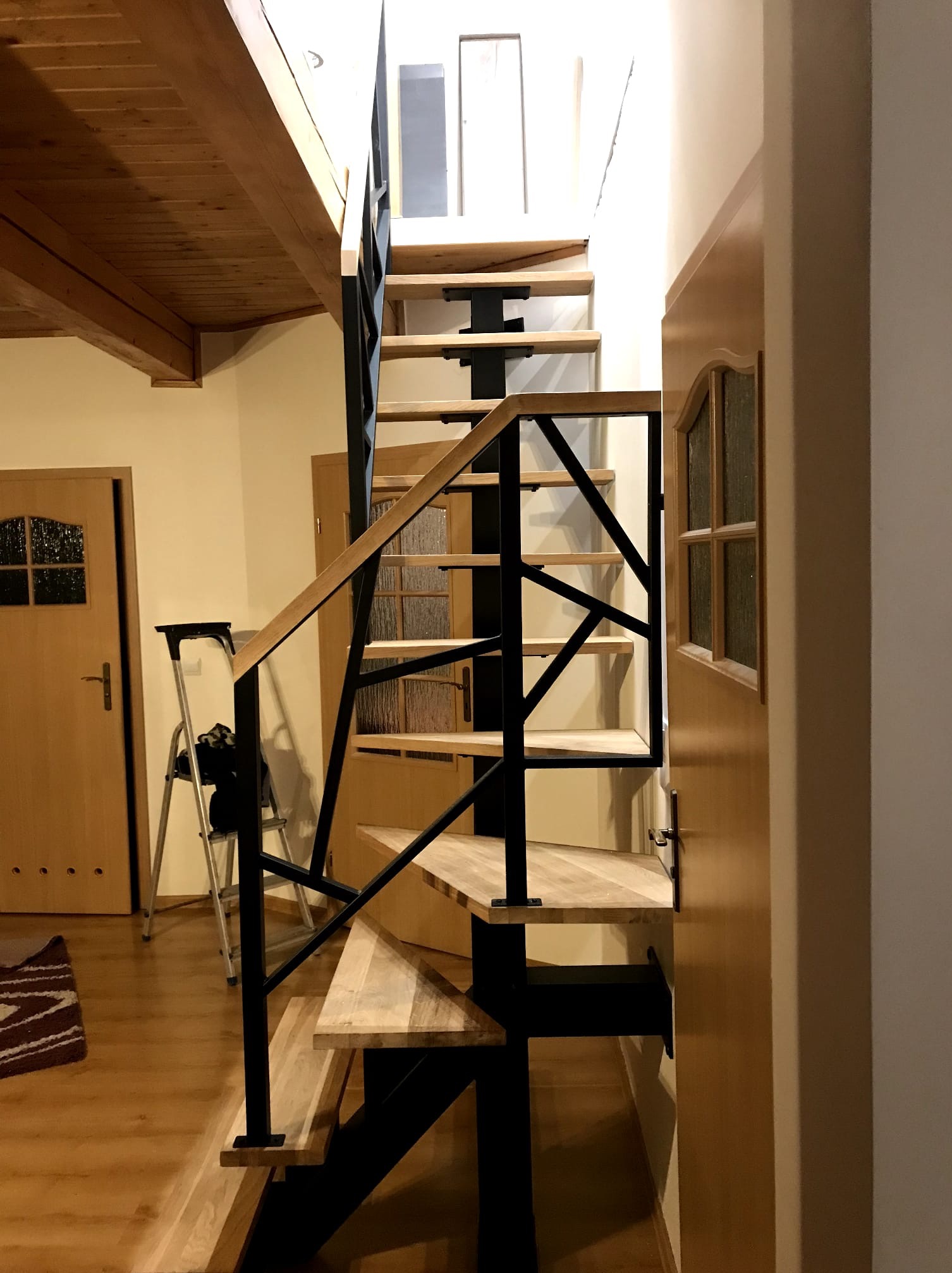 Nowoczesne schody drewniane z czarną metalową balustradą w jasnym wnętrzu. Widoczne drewniane drzwi i sufit. Na podłodze leży dywanik.