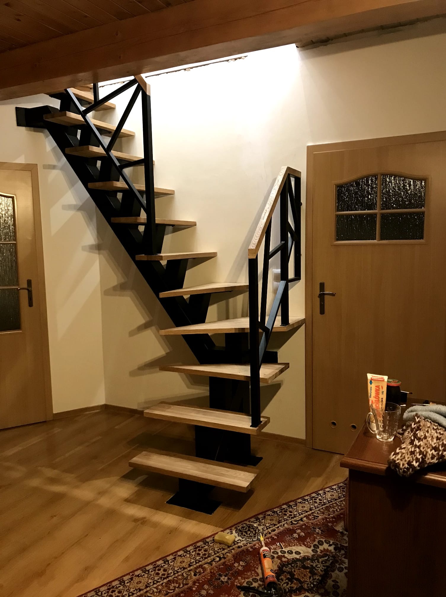 Nowoczesne schody drewniane z czarną metalową konstrukcją, zamontowane w jasnym wnętrzu, widoczne drzwi i fragment komody. Minimalistyczny design.